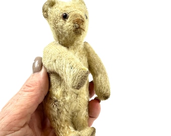 May include: A small, vintage-style teddy bear with worn, cream-colored fur, dark brown bead eyes, and a stitched nose. The bear features articulated limbs, rounded ears, and a textured, aged appearance throughout its entire body structure.