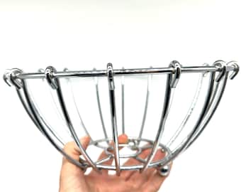 Può includere: Una ciotola per frutta in filo metallico color argento costruita con sottili aste di metallo curve che formano una struttura arrotondata a telaio aperto. La ciotola presenta una base circolare e una serie di fili verticali verso un bordo superiore.