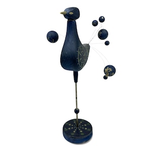 May include: A dark blue wooden bird figurine standing on a circular base. The bird features a long metal neck, a painted body with green dot patterns, and a tail made of thin metal wires tipped with small blue spheres decorated with gold accents.