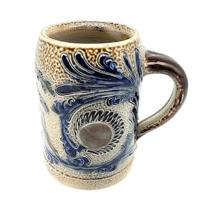 Puede incluir: Una jarra de cerveza de gres esmaltado con sal que presenta un cuerpo beige texturizado con un acabado moteado. La jarra est&aacute; decorada con patrones florales y de volutas en relieve azul cobalto que rodean un medall&oacute;n circular central.