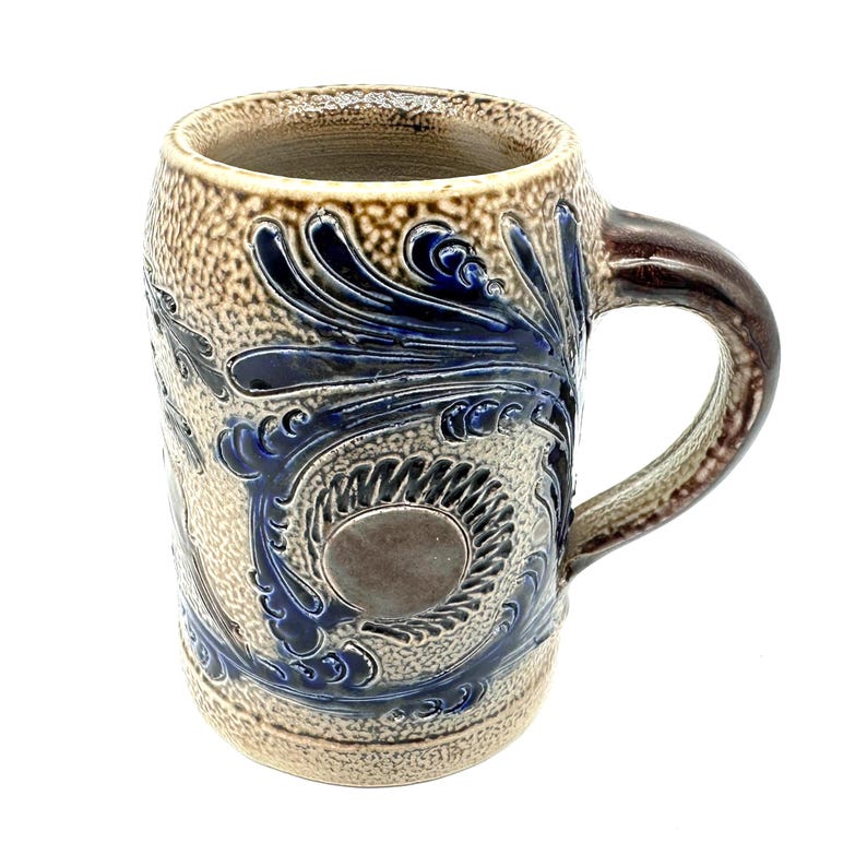 Puede incluir: Una jarra de cerveza de gres esmaltado con sal que presenta un cuerpo beige texturizado con un acabado moteado. La jarra est&aacute; decorada con patrones florales y de volutas en relieve azul cobalto que rodean un medall&oacute;n circular central.