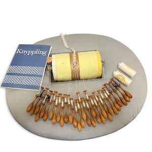 May include: A bobbin lace making kit featuring a yellow cylindrical pillow, numerous wooden bobbins with white thread, two spools of thread, and a blue instructional book titled Knyppling by Sally Johansson, all arranged on a gray surface.