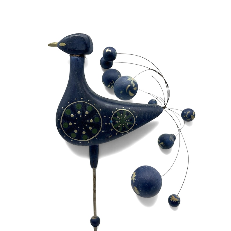 May include: A dark blue wooden bird garden stake featuring a painted green and white circular pattern on its body. The bird has a gold-colored beak and a decorative tail made of thin metal wires tipped with small, round, dark blue wooden spheres.
