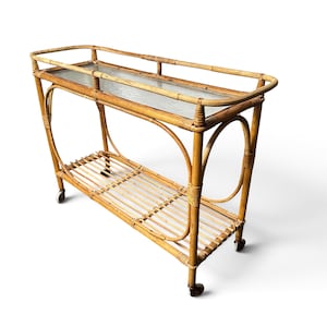 Può includere: Un carrello di servizio rettangolare in rattan con finitura naturale color marrone chiaro, un ripiano superiore con inserto in vetro e un ripiano inferiore a doghe. La struttura è sostenuta da quattro piccole ruote e presenta supporti laterali curvi.