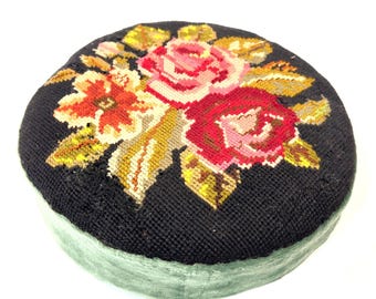 May include: A round, flat cushion featuring a black needlepoint top with a floral design of pink and red roses surrounded by green leaves. The sides of the cushion are covered in a solid, light green fabric with a soft, velvet-like texture.