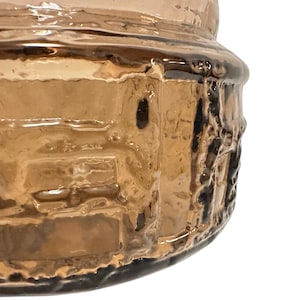 May include: A cylindrical glass container with a warm amber hue, featuring a textured surface with raised vertical and horizontal ridges that create a grid-like pattern across the exterior of the vessel, showing a smooth rim at the top.