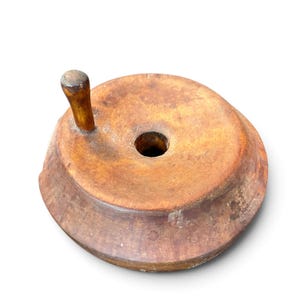 May include: A circular wooden object with a natural brown finish, featuring a central hole and a small, upright handle attached to the top surface. The piece has a tapered, sloped edge and shows visible wood grain and texture throughout its surface.