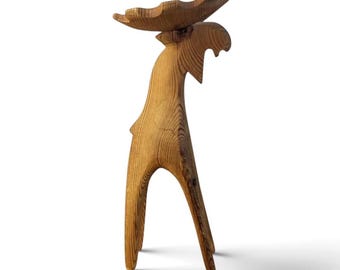 May include: A standing wooden moose figurine crafted from light brown wood with a visible grain pattern. The sculpture features a long neck, two slender legs, and a wide, flat set of antlers extending horizontally from the top of the head.