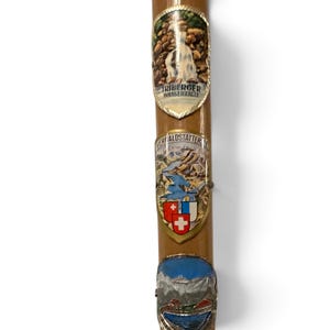 Puede incluir: Un bast&oacute;n de madera de color marr&oacute;n claro adornado con tres placas met&aacute;licas de recuerdo. La placa superior representa las Triberger Wasserf&auml;lle, la placa central muestra el Vierwaldst&auml;ttersee con un escudo y la placa inferior presenta un paisaje monta&ntilde;oso.