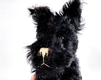 Puede incluir: Un perro de peluche con pelaje negro largo y desgreñado, orejas puntiagudas erguidas y un detalle de nariz y boca bordado en color beige. El animal de peluche presenta ojos oscuros y brillantes incrustados en el denso material negro.