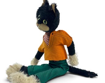 Puede incluir: Un muñeco de gato de peluche con pelaje negro, hocico color crema y ojos verdes. El muñeco viste una camiseta naranja de manga corta con cuello a rayas rojas y blancas, pantalones verdes y tiene pelaje largo y desgreñado color crema.