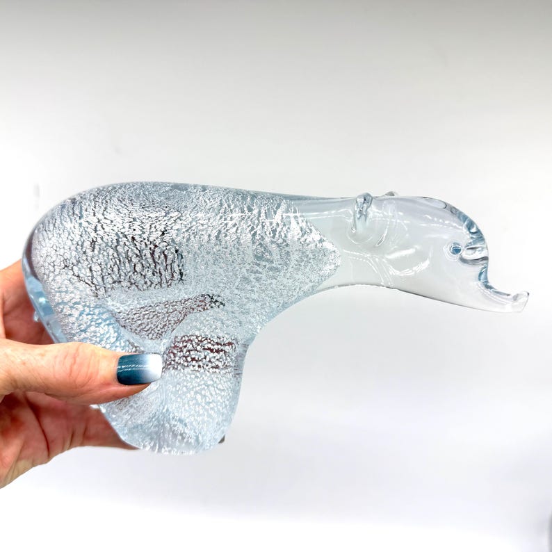 May include: A clear glass figurine shaped like a polar bear, featuring a textured, silver-flecked interior that creates a shimmering effect throughout the body, while the head and neck remain smooth and transparent glass.