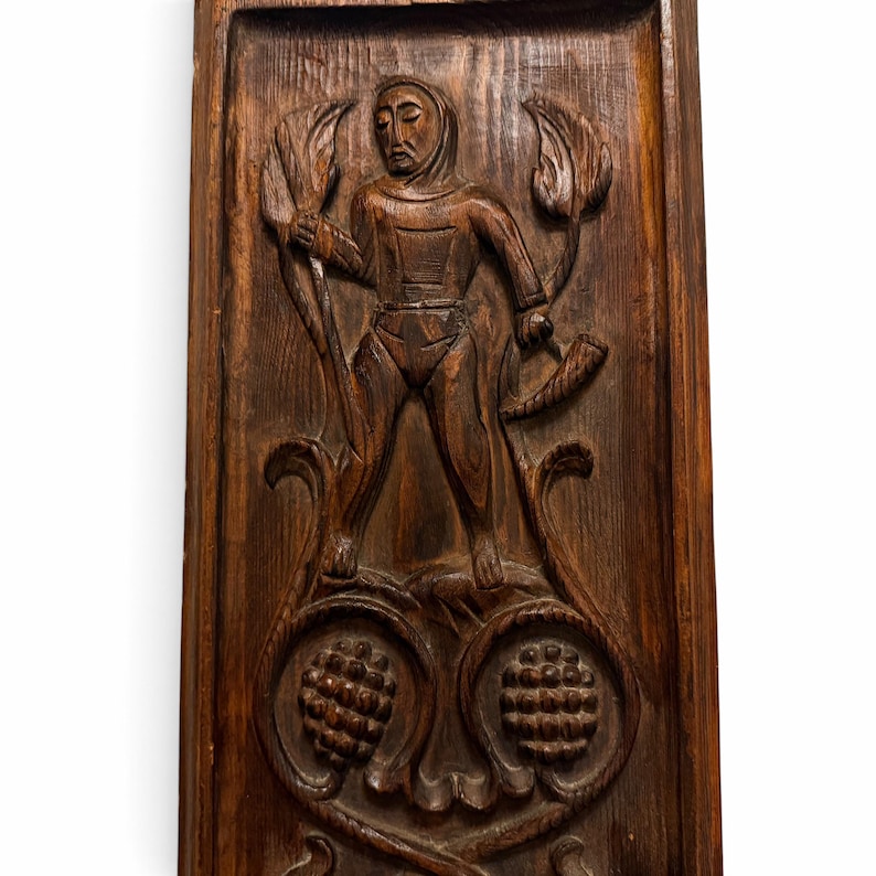 May include: A rectangular dark brown wooden wall panel featuring a hand-carved relief of a figure wearing a hood. The figure holds a staff and a curved horn, surrounded by stylized leaf motifs and two prominent pinecone carvings at the base.