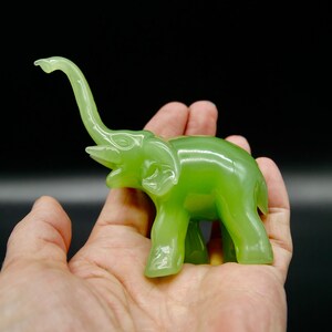 K&ouml;nnte beinhalten: Eine kleine, jadegr&uuml;ne Elefantenfigur mit erhobenem R&uuml;ssel und detaillierten Merkmalen. Die Figur wird in einer Hand vor dunklem Hintergrund gehalten, wodurch ihre glatte, polierte Oberfl&auml;che zur Geltung kommt. Der R&uuml;ssel des Elefanten ist nach oben gebogen.