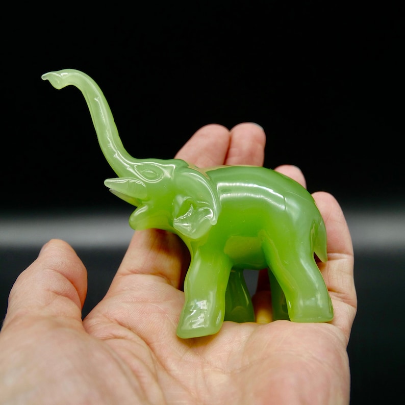 K&ouml;nnte beinhalten: Eine kleine, jadegr&uuml;ne Elefantenfigur mit erhobenem R&uuml;ssel und detaillierten Merkmalen. Die Figur wird in einer Hand vor dunklem Hintergrund gehalten, wodurch ihre glatte, polierte Oberfl&auml;che zur Geltung kommt. Der R&uuml;ssel des Elefanten ist nach oben gebogen.