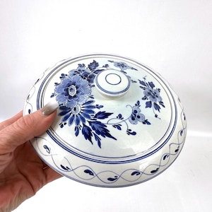 May include: A white ceramic serving dish lid with blue floral patterns. The lid has a central knob and a repeating design around the edge. The floral design includes large flowers and leaves.