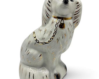 Puede incluir: Una figura de perro de cerámica blanca en posición sentada, con detalles pintados en oro sobre el pelaje, un collar dorado con una pequeña campana colgante y un detalle de cadena dorada sobre el pecho. El perro tiene ojos y nariz pintados de negro.