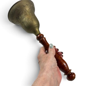 May include: A hand bell featuring a polished brass dome with an etched leaf pattern and a smooth, dark brown wooden handle. The bell has a flared shape and a rounded base on the handle, providing a clear contrast between the metal and wood.