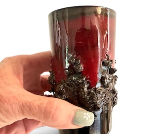 May include: A cylindrical ceramic tumbler featuring a glossy deep red glaze on the upper portion and a dark, heavily textured, volcanic-like black surface around the middle and base, creating a distinct contrast in both color and tactile finish.