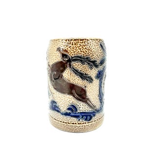 Puede incluir: Una taza cil&iacute;ndrica de cer&aacute;mica con un acabado texturizado de esmalte salino en color beige. El exterior presenta un motivo de ciervo saltando en marr&oacute;n oscuro y patrones de olas en azul profundo en la base, con est&eacute;tica de gres.