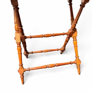 Puede incluir: Un soporte plegable de madera con cuatro patas torneadas en un acabado marr&oacute;n c&aacute;lido. La estructura incluye dos travesa&ntilde;os horizontales con detalles de husillo decorativos que conectan las patas para dar estabilidad al marco.