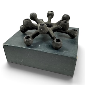 Puede incluir: Un candelabro negro mate de múltiples brazos con una superficie texturizada y granulada. El objeto presenta un anillo central conectado a ocho brazos radiantes, cada uno terminado en una copa circular para velas. Descansa sobre una base rectangular de color gris oscuro.