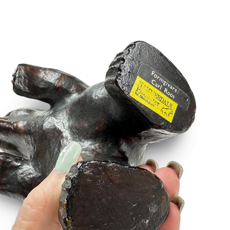 May include: A dark brown, textured bear figurine with a yellow rectangular label on the base. The label features black text reading Formgivare: Carl Roos, H&Auml;RJEDALS, PRODUKTER, and includes a small bear icon and a telephone number.