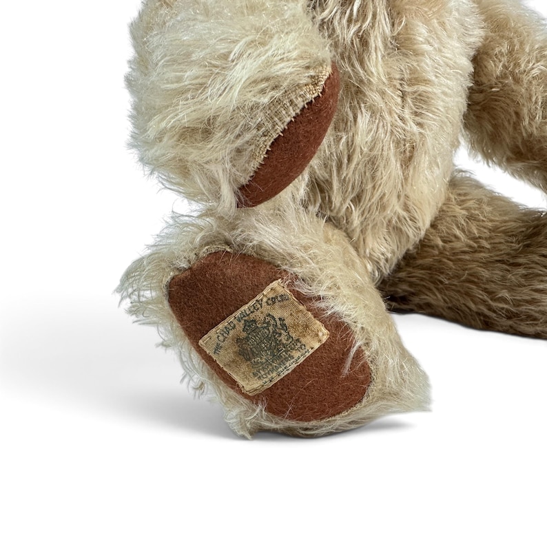 K&ouml;nnte beinhalten: Ein beigefarbener Pl&uuml;schteddyb&auml;r mit langem, zotteligem Fell. Der B&auml;r hat dunkelbraune Filzpfoten. Ein rechteckiges Stoffetikett ist an der Unterseite eines Fu&szlig;es angen&auml;ht und zeigt den Text "CHAD VALLEY" sowie ein Wappenlogo in schwarzer Tinte.