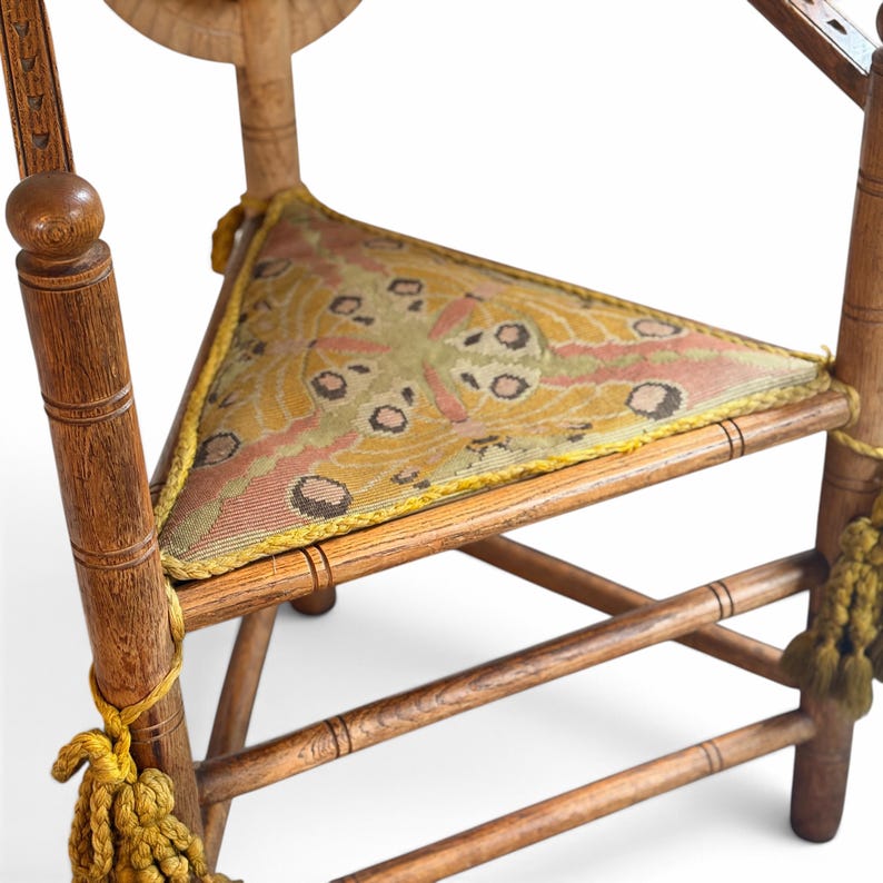 May include: A wooden chair featuring a triangular seat upholstered in a patterned fabric with yellow, pink, and dark brown tones. The chair has turned wooden legs and arm supports, accented with decorative yellow tassels hanging from the frame.