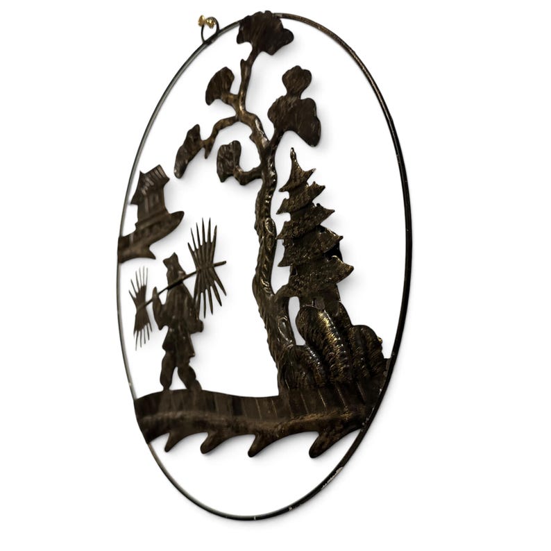 May include: A dark bronze-colored oval metal wall art piece featuring a cutout silhouette of a person carrying a pole with bundles, a tall tree, a multi-tiered pagoda structure, and a small building, all enclosed within a thin circular frame.