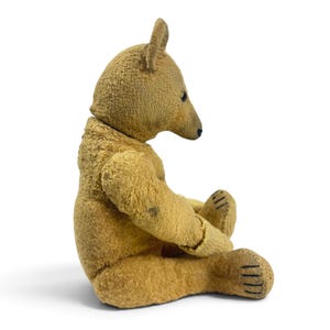 Pode incluir: Um urso de peluche de estilo vintage feito de tecido castanho-claro texturizado. O urso &eacute; mostrado numa posi&ccedil;&atilde;o sentada de perfil, com um focinho pontiagudo, olhos pequenos e escuros, um nariz cosido e detalhes de fio escuro nas patas.