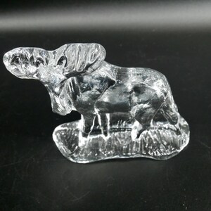 May include: A clear glass sculpture of a ram, standing on a textured base. The ram is detailed with visible features, including horns and fur. The sculpture is set against a dark background.