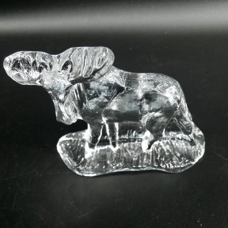 May include: A clear glass sculpture of a ram, standing on a textured base. The ram is detailed with visible features, including horns and fur. The sculpture is set against a dark background.