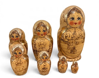 Puede incluir: Un conjunto de seis muñecas rusas de madera con rostros pintados a mano, pestañas detalladas y labios rojos. Cada muñeca presenta un acabado de madera clara con detalles dorados e ilustraciones en tinta negra de edificios.