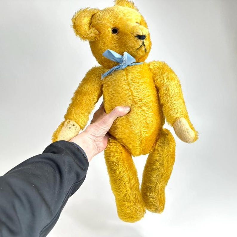 May include: A vintage golden yellow teddy bear with a blue ribbon tied around its neck. The bear has black eyes and a black stitched nose. Its arms and legs are slightly worn, and it is being held by a person.