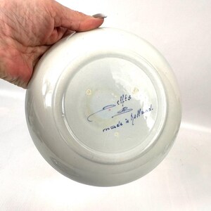May include: A white ceramic plate with a blue and white design. The plate has a slightly curved rim and a smooth surface. The bottom of the plate has blue handwritten text that reads "elfts made in Holland."