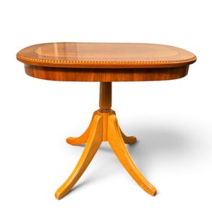 Op de afbeelding: Een houten zuiltafel met een ovaal blad met een glad, gepolijst oppervlak en een decoratieve kralenrand. De tafel wordt ondersteund door een centrale kolom die zich vertakt in drie gebogen poten, alles afgewerkt in een warme, lichtbruine tint.
