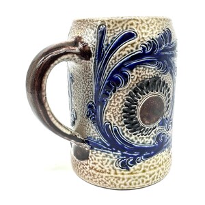 Puede incluir: Una jarra de cerveza de cer&aacute;mica con una superficie beige texturizada y moteada, y un asa de color marr&oacute;n oscuro. La jarra presenta patrones en relieve de color azul cobalto con hojas de acanto y un medall&oacute;n circular central.