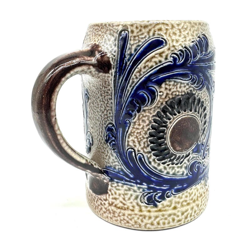 Puede incluir: Una jarra de cerveza de cer&aacute;mica con una superficie beige texturizada y moteada, y un asa de color marr&oacute;n oscuro. La jarra presenta patrones en relieve de color azul cobalto con hojas de acanto y un medall&oacute;n circular central.