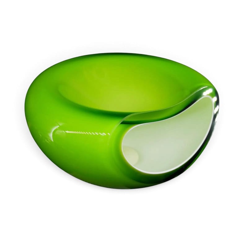May include: A smooth, rounded glass bowl featuring a vibrant lime green exterior and a contrasting opaque white interior. The vessel has a wide, open top and a curved, asymmetrical side cutout that reveals the white inner surface of the glass object.