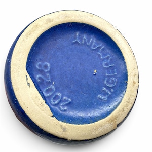 May include: The circular base of a ceramic object features a deep blue glazed center with embossed text reading 200 28 and W. GERMANY. A wide, unglazed, off-white circular rim frames the blue center, showing a textured, matte finish.