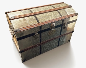 Op de afbeelding: Een rechthoekige opbergkist met een gewelfd deksel, voorzien van een metallic zilveren afwerking met een gestructureerd patroon. De kist is verstevigd met donkerbruine houten latten, zwarte metalen banden en zilverkleurige metalen hoekbeschermers en sluitingen.