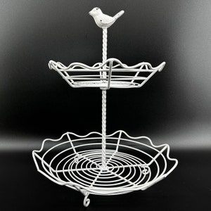 Könnte beinhalten: Ein weißer, zweistöckiger Servierständer mit einer Vogelfigur oben. Der Ständer hat eine gedrehte Mittelsäule und zwei Drahtkörbe. Das untere Tablett hat einen gewellten Rand, und der Ständer ist aus Metall.