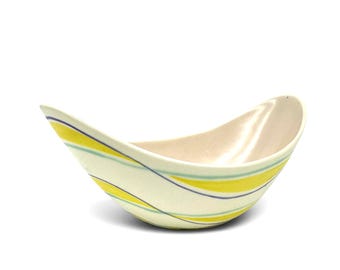 May include: A curved, boat-shaped ceramic bowl featuring an off-white interior and an exterior decorated with diagonal, flowing stripes in bright yellow, light blue, and thin dark grey lines, creating a dynamic and asymmetrical visual pattern.