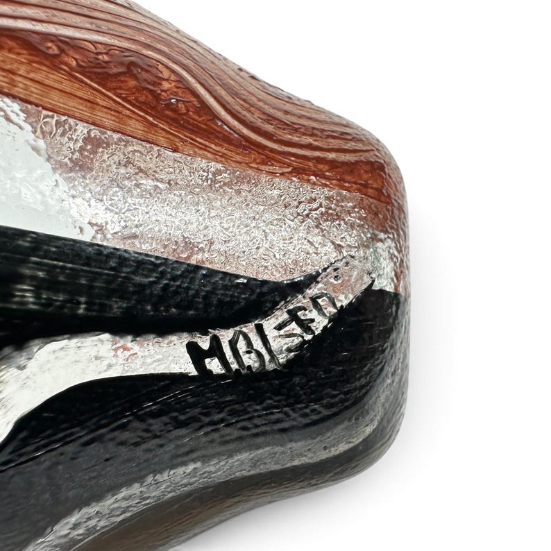 May include: A glass object featuring a swirling pattern of brown, black, and silver metallic tones. The surface has a textured, layered appearance with a glossy finish. The letters MBI 50 are clearly etched into the surface in black ink.