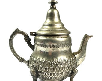 May include: A silver-toned metal teapot featuring a tall, pointed lid, a curved handle, and a spout. The body is decorated with a textured, repeating scale-like pattern around the base and a band of floral motifs, supported by three legs.