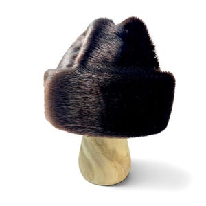 Op de afbeelding: Een donkerbruine bontmuts met een gestructureerde, ronde kroon en een brede, omgeslagen rand. De dichte vacht met korte haren heeft een glanzende textuur met subtiele kleurvariaties, variërend van diep espresso tot lichtere chocoladebruine tinten.