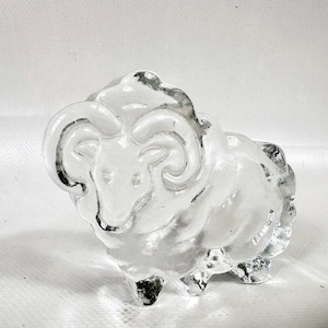 May include: A clear glass ram ornament with detailed horns and a textured body. The ram stands on four legs and has a slightly transparent appearance, creating a unique decorative piece.