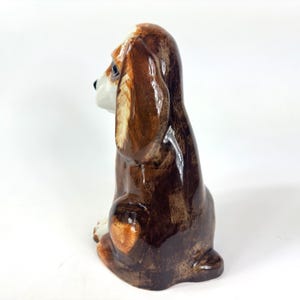 Peut inclure: Figurine en c&eacute;ramique d'un chien beagle assis. Le chien a des poils bruns et blancs avec de longues oreilles tombantes. La figurine est brillante et d&eacute;taill&eacute;e, avec une apparence r&eacute;aliste. Le chien est en position assise.