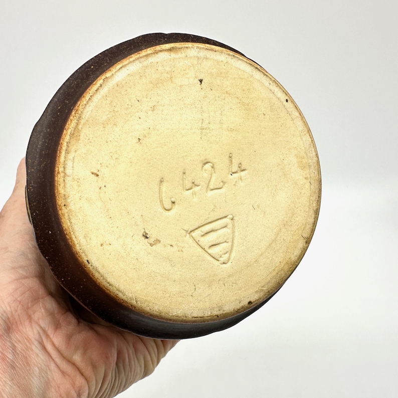 Peut inclure: Vue du dessous d'un vase en c&eacute;ramique marron. La base est de couleur beige clair avec le num&eacute;ro "6424" et un symbole triangulaire grav&eacute; sur la surface. Le vase a un ext&eacute;rieur brun fonc&eacute; textur&eacute;.